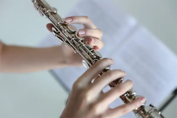 Gros plan sur les mains d'une personne jouant d'une flûte traversière argentée. Les doigts sont délicatement posés sur les touches et les boutons de l'instrument. On aperçoit une partie du corps de la flûte, brillante et dont les mécanismes sont clairement visibles. À l'arrière-plan, légèrement flou, on aperçoit un pupitre avec une partition de musique. La lumière est douce et diffuse, se concentrant sur les mains et l'instrument de musique.